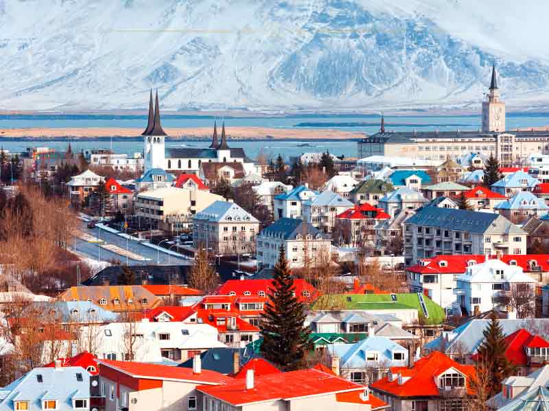 A small town with small houses, on the shores of the sea. The houses have red rooftops. On the other side of the water there is a large mountain covered with snow.